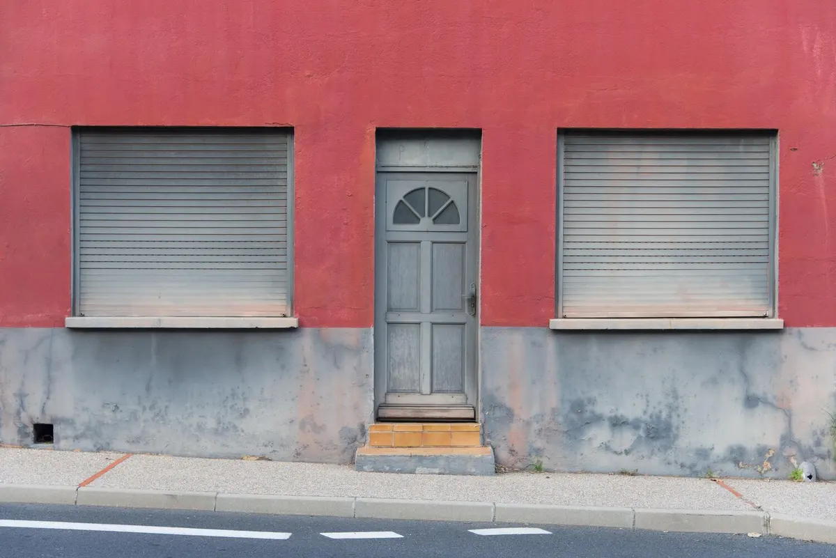 Deux volets roulants à lames grises baissés de part et d'autre d'une porte en bois, sur une façade enduite rouge au soubassement gris